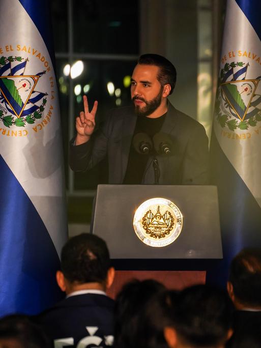 Nayib Bukele steht an einem Rednerpult und spricht zum Publikum. Links und rechts von ihm hängt die blau-weiße Flagge von El Salvador.