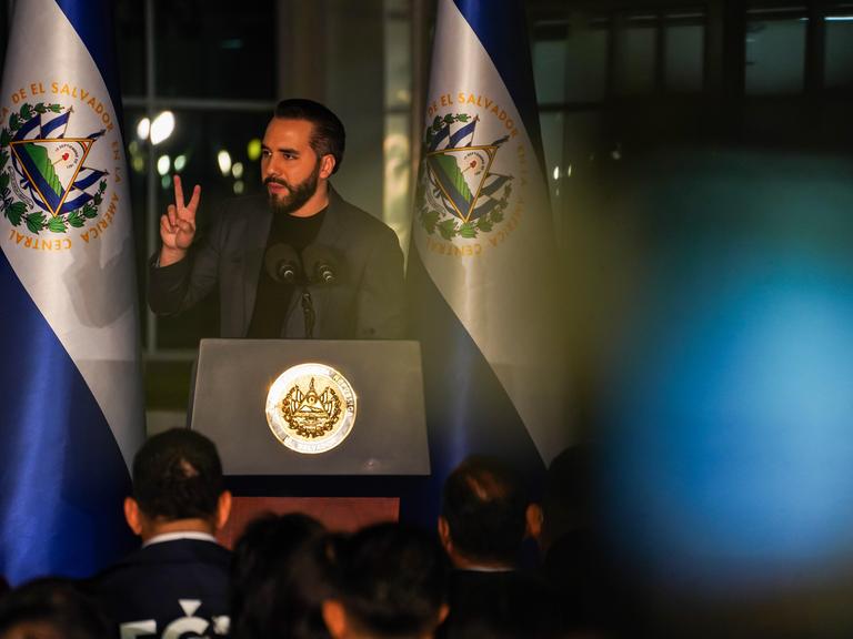 Nayib Bukele steht an einem Rednerpult und spricht zum Publikum. Links und rechts von ihm hängt die blau-weiße Flagge von El Salvador.