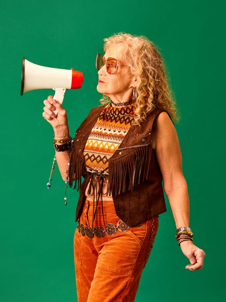 Eine extravagante ältere Frau mit langen lockigen rotblonden Haaren und Hippie-Outfit hält ein Megaphon vor den Mund.