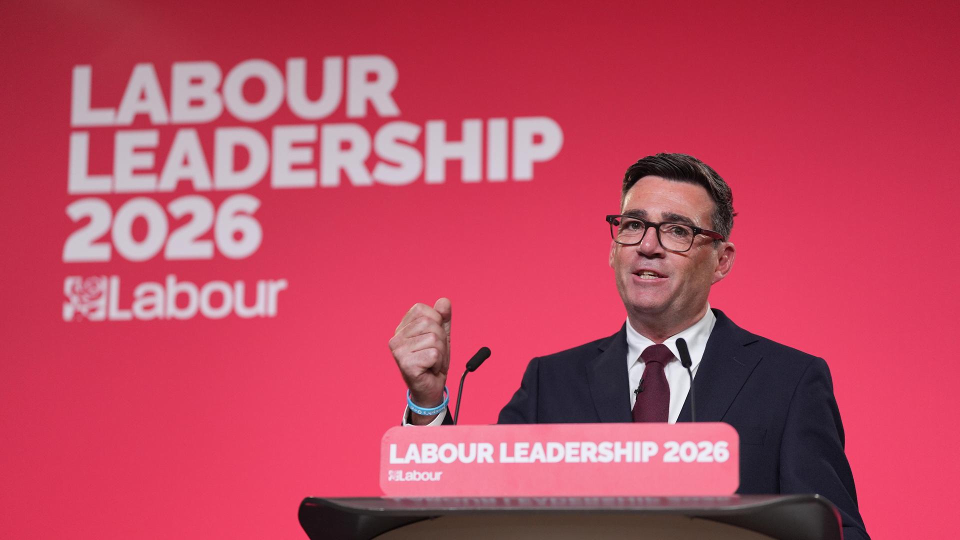 Burnham steht an einem Rednerpult, ballt die rechte Faust und spricht in ein Mikrofon. Auf einer roten Wand hinter ihm steht in weißer Schrift "Labour Leadership 2026"