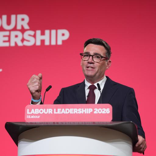 Burnham steht an einem Rednerpult, ballt die rechte Faust und spricht in ein Mikrofon. Auf einer roten Wand hinter ihm steht in weißer Schrift "Labour Leadership 2026"