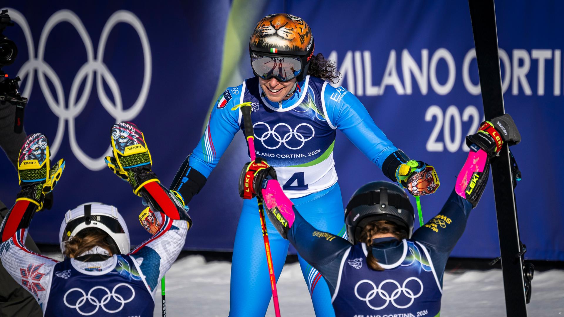 Man sieht die beiden im Schnee knieenden Gratulantinnen von hinten. Die lachende Brignone im blauen Renndress sieht man von vorne. Dahinter eine blaue Plane mit weißer Schrift und den Olympischen Ringen.