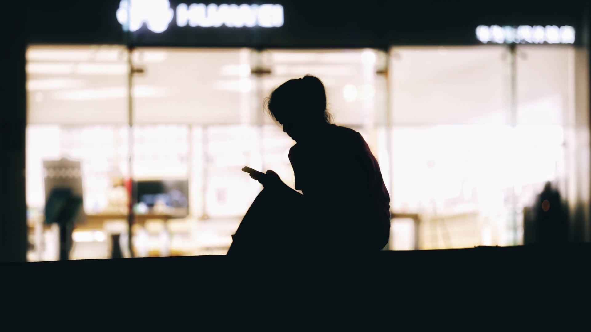 Die Silhouette einer Frau, die vor einer beleuchteten Scheibe sitzt und auf ihr Smartphone schaut. 