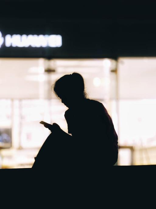 Die Silhouette einer Frau, die vor einer beleuchteten Scheibe sitzt und auf ihr Smartphone schaut. 