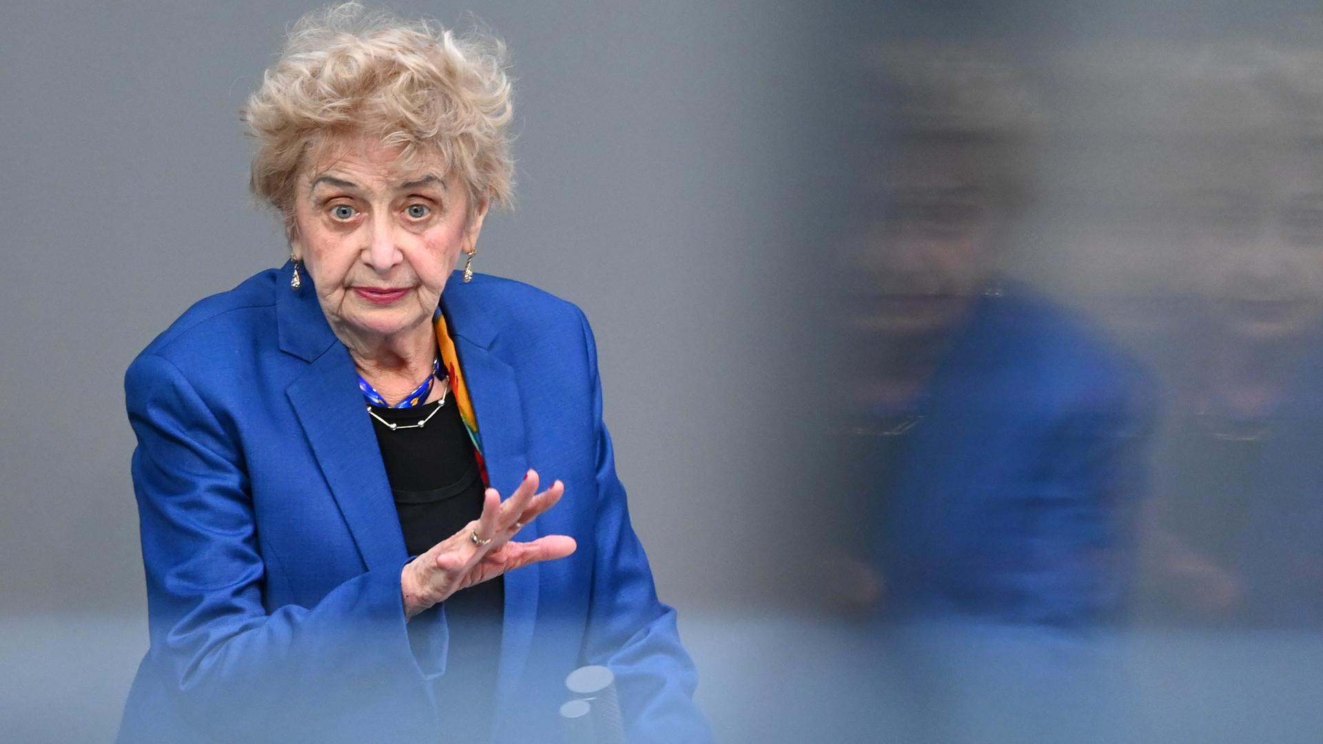 Friedman trägt ein blaues Jackett. Sie spricht und gestikuliert mit beiden Händen. Der Hintergrund ist unscharf, sie spiegelt sich in einer Glasscheibe. 