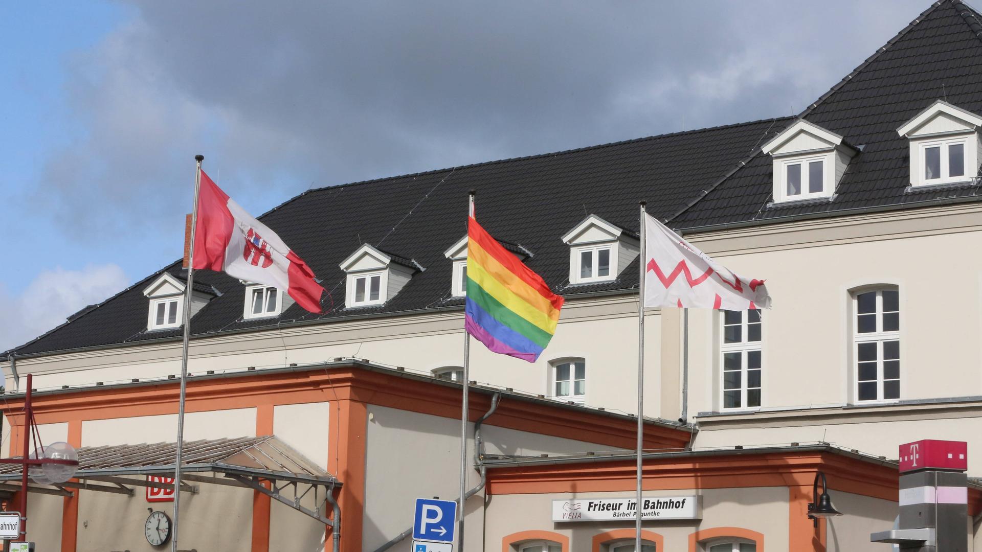 Drei Flaggen auf einem Fahnenmast vor einem weißen Gebäude in Neubrandenburg. In der Mitte die Regenbogenflagge. Links davon die Flagge Neubrandenburgs.