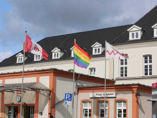Drei Flaggen auf einem Fahnenmast vor einem weißen Gebäude in Neubrandenburg. In der Mitte die Regenbogenflagge. Links davon die Flagge Neubrandenburgs.