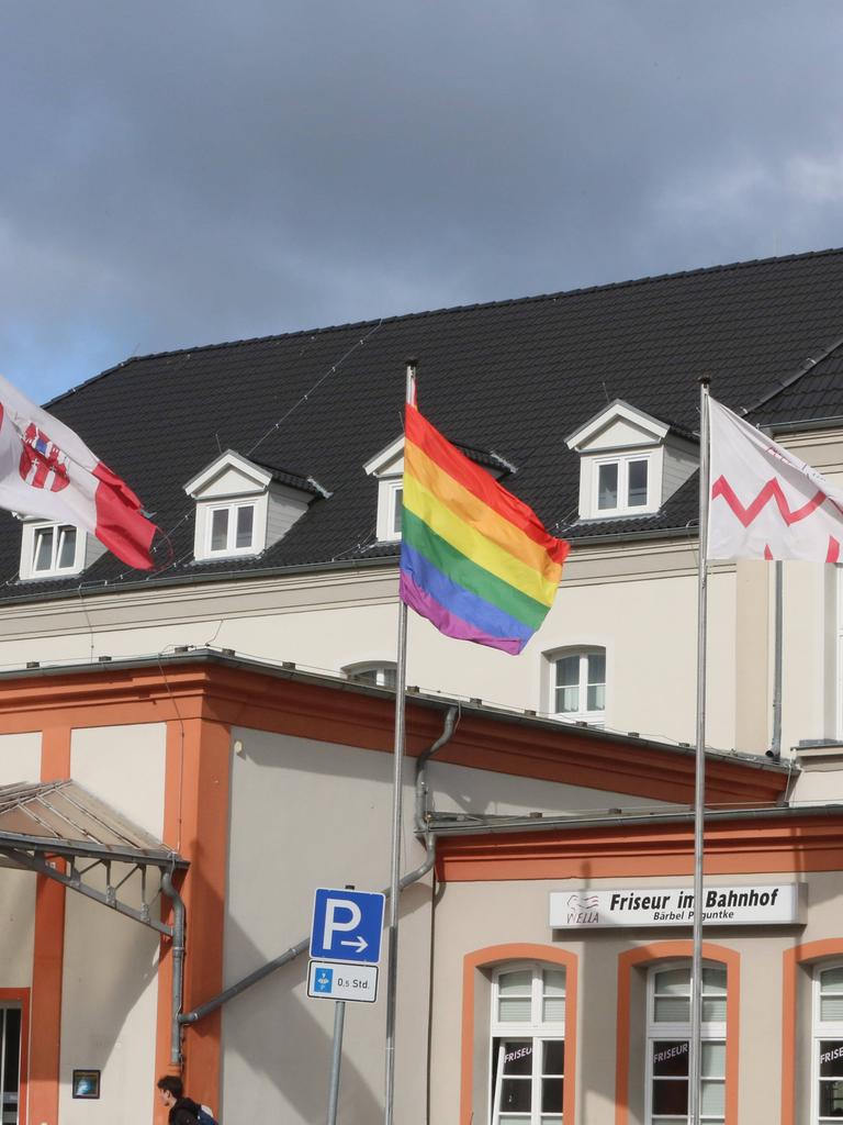 Drei Flaggen auf einem Fahnenmast vor einem weißen Gebäude in Neubrandenburg. In der Mitte die Regenbogenflagge. Links davon die Flagge Neubrandenburgs.