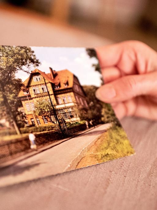 Eine Frau, die als Kind in die Kinderheilanstalt „Waldhaus“ Bad Salzdetfurth geschickt wurde, hält in ihrer Wohnung eine historische Postkarte Anstalt, in der es in der Nachkriegszeit zu schweren Misshandlungen von Klein- und Schulkindern kam. 
