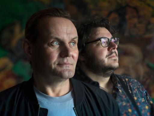 Devid Striesow und Axel Ranisch stehen in einem dunklen Raum und schauen zur Seite.