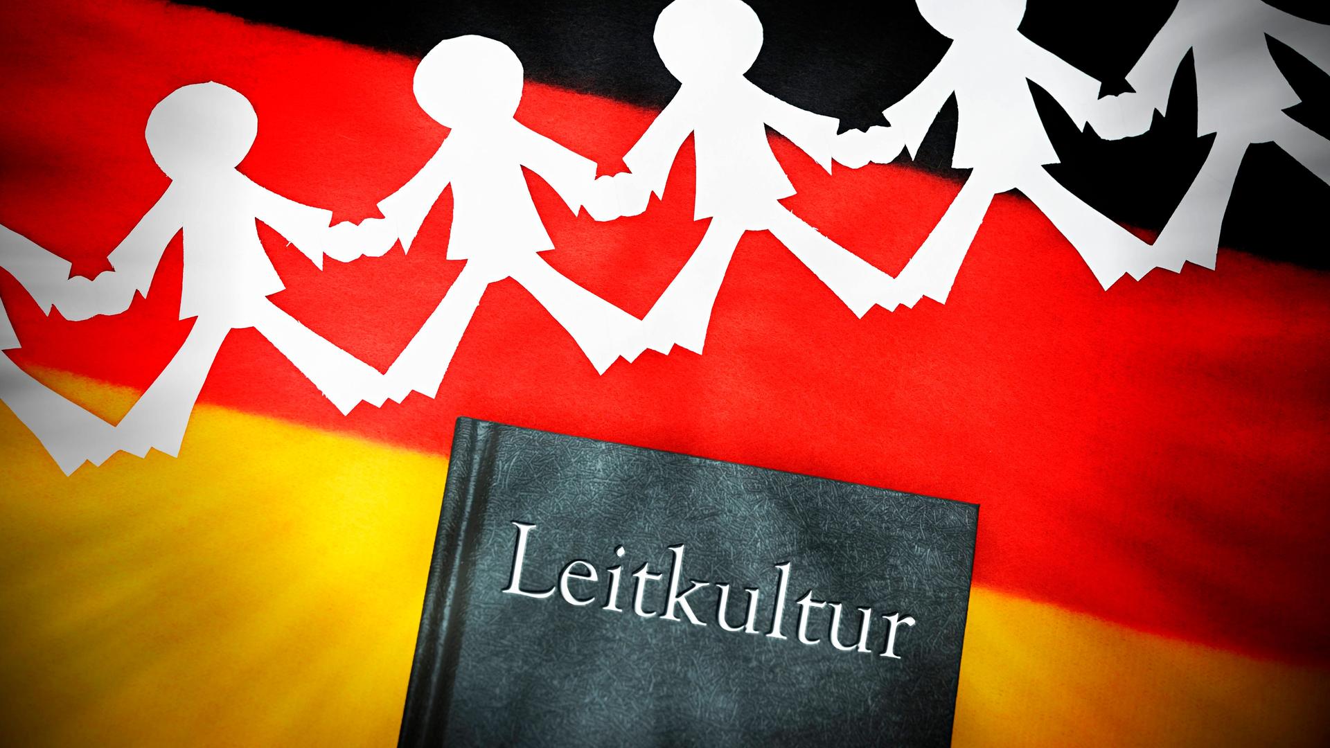 Ein schwarzes Buch mit dem silber gedruckten Wort "Leitkultur" auf einer deutschen Flagge. Über dem Buch liegt ein weißer Scherenschnitt, der Menschen darstellt, die sich an den Händen halten. 