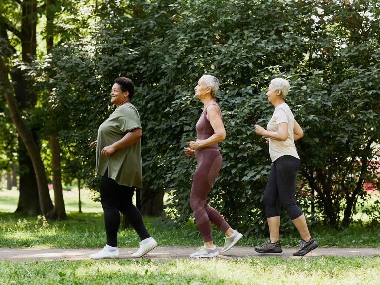 Drei ältere Frauen joggen fröhlich im Park, sie laufen hintereinander in einer Reihe.
