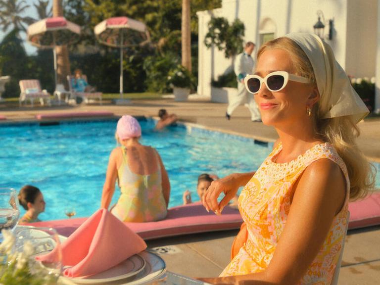 Im Still aus "Palm Royale" sitzt Kristen Wiig in einem Club an einem Pool.