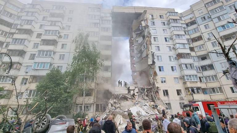 Ein Wohnhaus in einem Häuserblock ist in der Mitte eingestürzt. Mehrere Menschen schauen von unten auf die Trümmer.