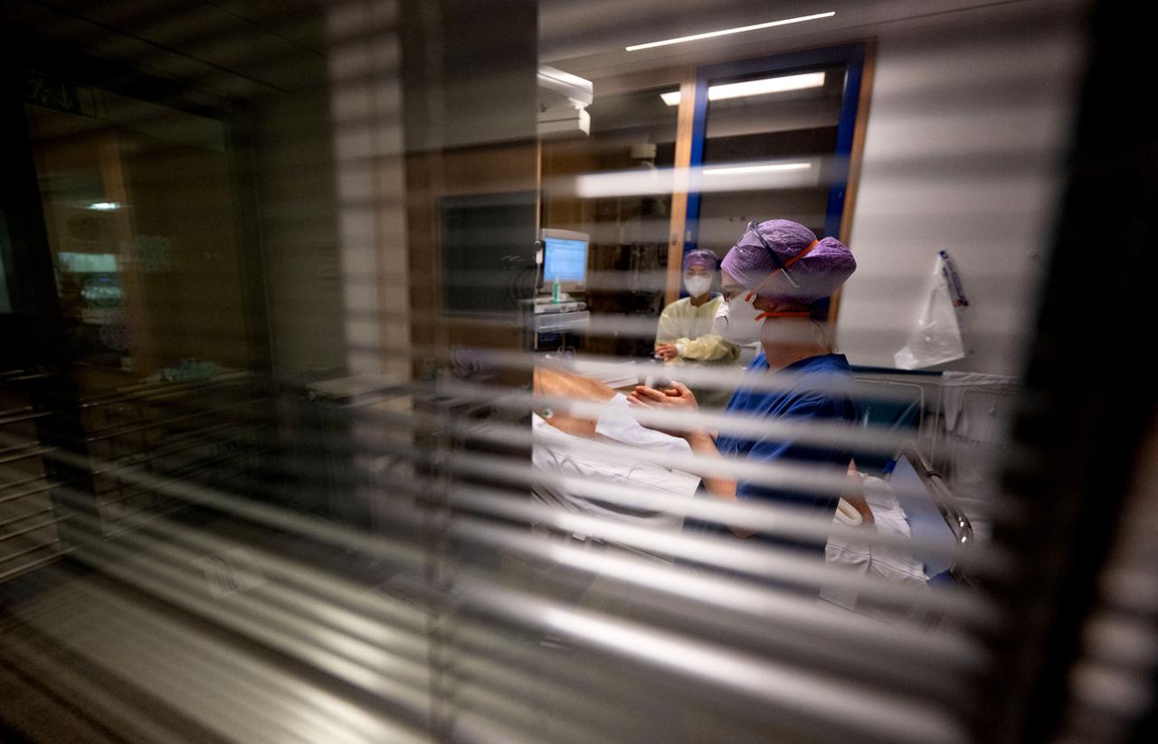 Durch ein Fenster mit halb geschlossenen Jalousien werden Krankenschwestern beobachtet, die sich auf der Intensivstation um einen Komapatienten kümmern.