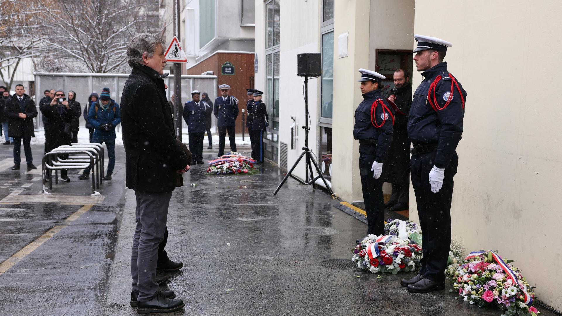 Der französische Karikaturist gedenkt der Opfer des Terroranschlags auf Charlie Hebdo. Vor ihm stehen zwei Polizisten. Auf dem Boden liegen Kränze. 