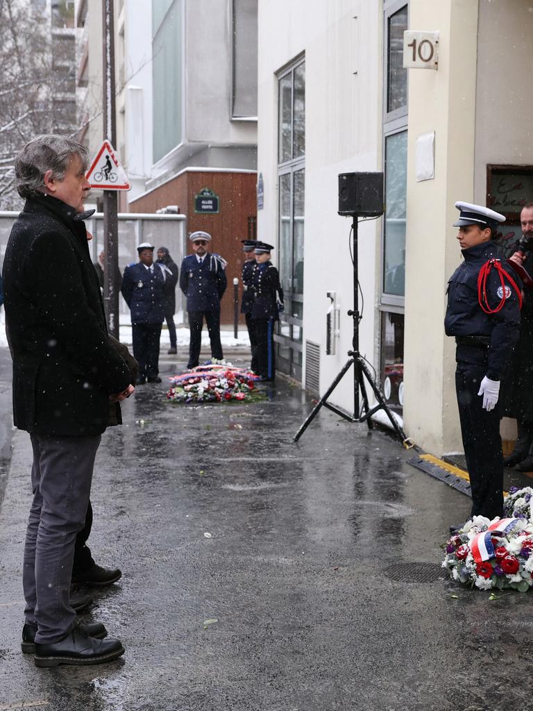 Der französische Karikaturist gedenkt der Opfer des Terroranschlags auf Charlie Hebdo. Vor ihm stehen zwei Polizisten. Auf dem Boden liegen Kränze. 
