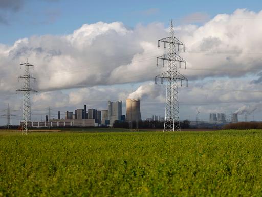 Impression vom RWE-Kraftwerk Bergheim Niederaußem hinter eine gründen Wiese. Das Kohlekraftwerk zieht seine Schwaden in die kalte Winterluft. In der Umgebung glänzen Strommasten, Windräder und Wolkenformationen in der kalten Wintersonne. 