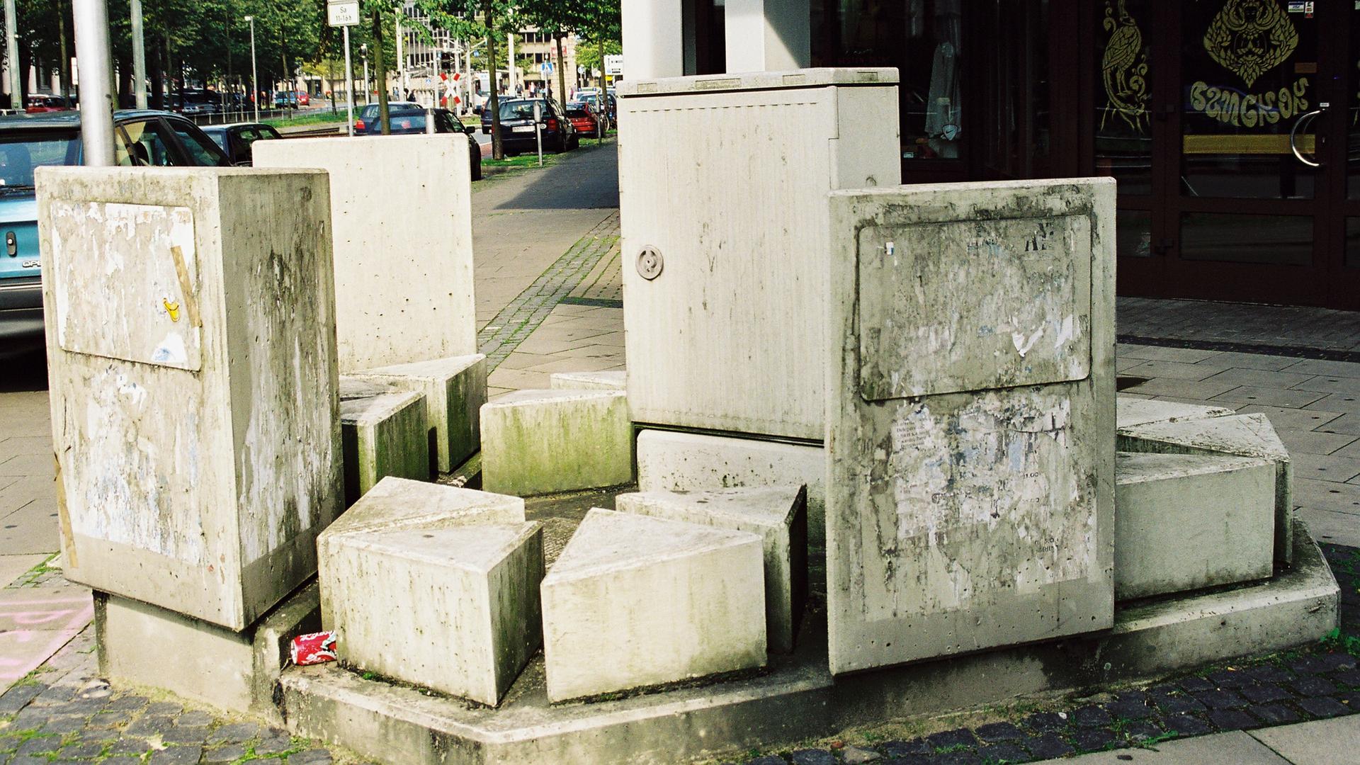 Stromkasten in Bielefeld, umgeben von mehreren unterschiedlich hohen Stelen, im Hintergrund die Straße 