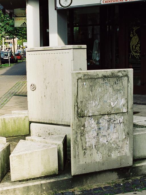 Stromkasten in Bielefeld, umgeben von mehreren unterschiedlich hohen Stelen, im Hintergrund die Straße 