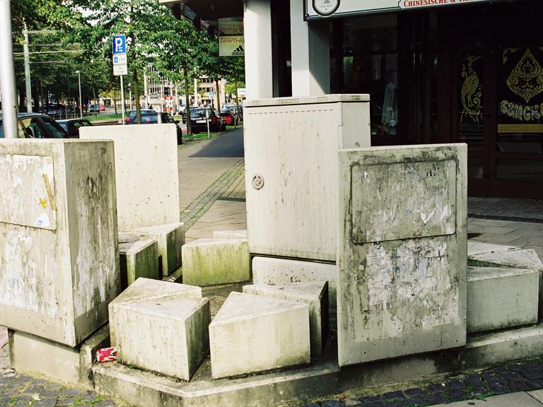 Stromkasten in Bielefeld, umgeben von mehreren unterschiedlich hohen Stelen, im Hintergrund die Straße 