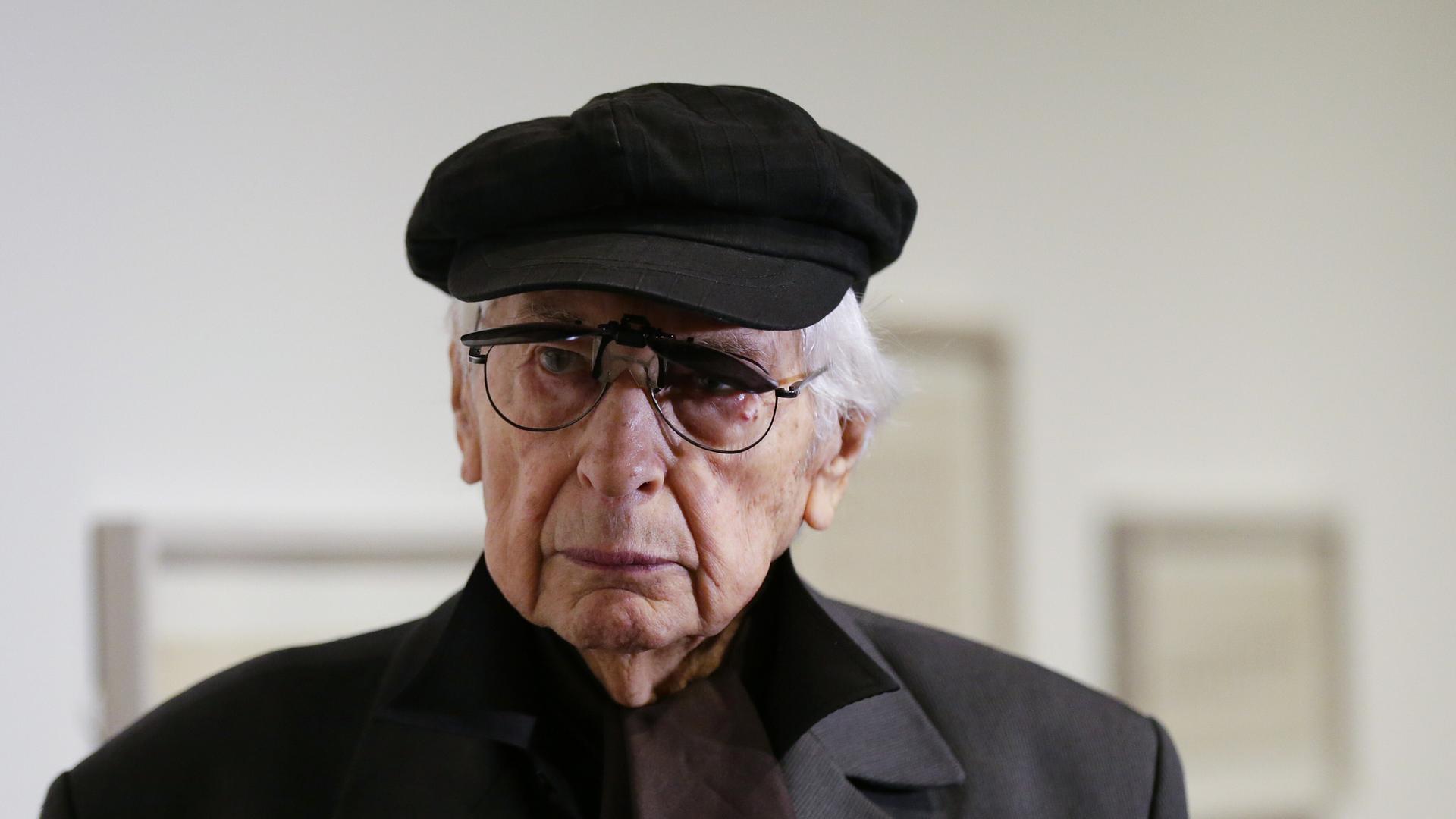 Der argentinische Künstler Julio Le Parc trägt Mütze und Mantel. Im Hintergrund sind unscharf Gemälde zu erkennen. Der argentinische Künstler Julio Le Parc trägt Mütze und Mantel. Im Hintergrund sind unscharf Gemälde zu erkennen.