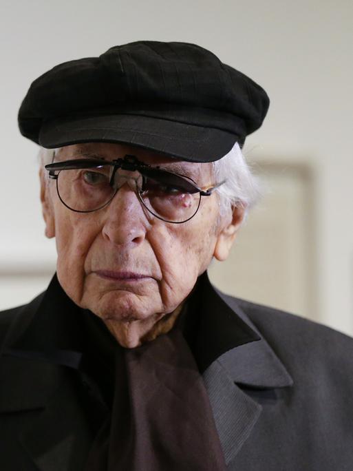 Der argentinische Künstler Julio Le Parc trägt Mütze und Mantel. Im Hintergrund sind unscharf Gemälde zu erkennen.
