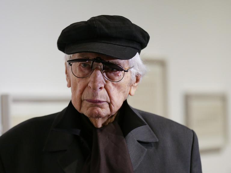 Der argentinische Künstler Julio Le Parc trägt Mütze und Mantel. Im Hintergrund sind unscharf Gemälde zu erkennen.