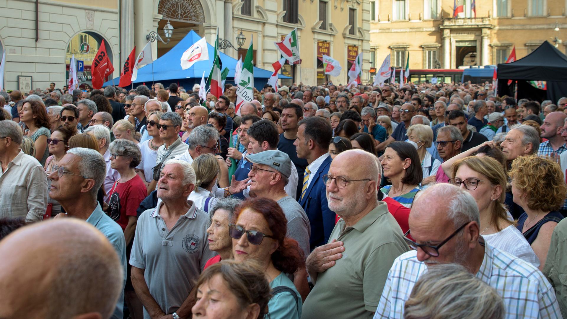 Menschen stehen dicht gedrängt auf einem Platz in Rom, manche halten Fahnen oder Transparente