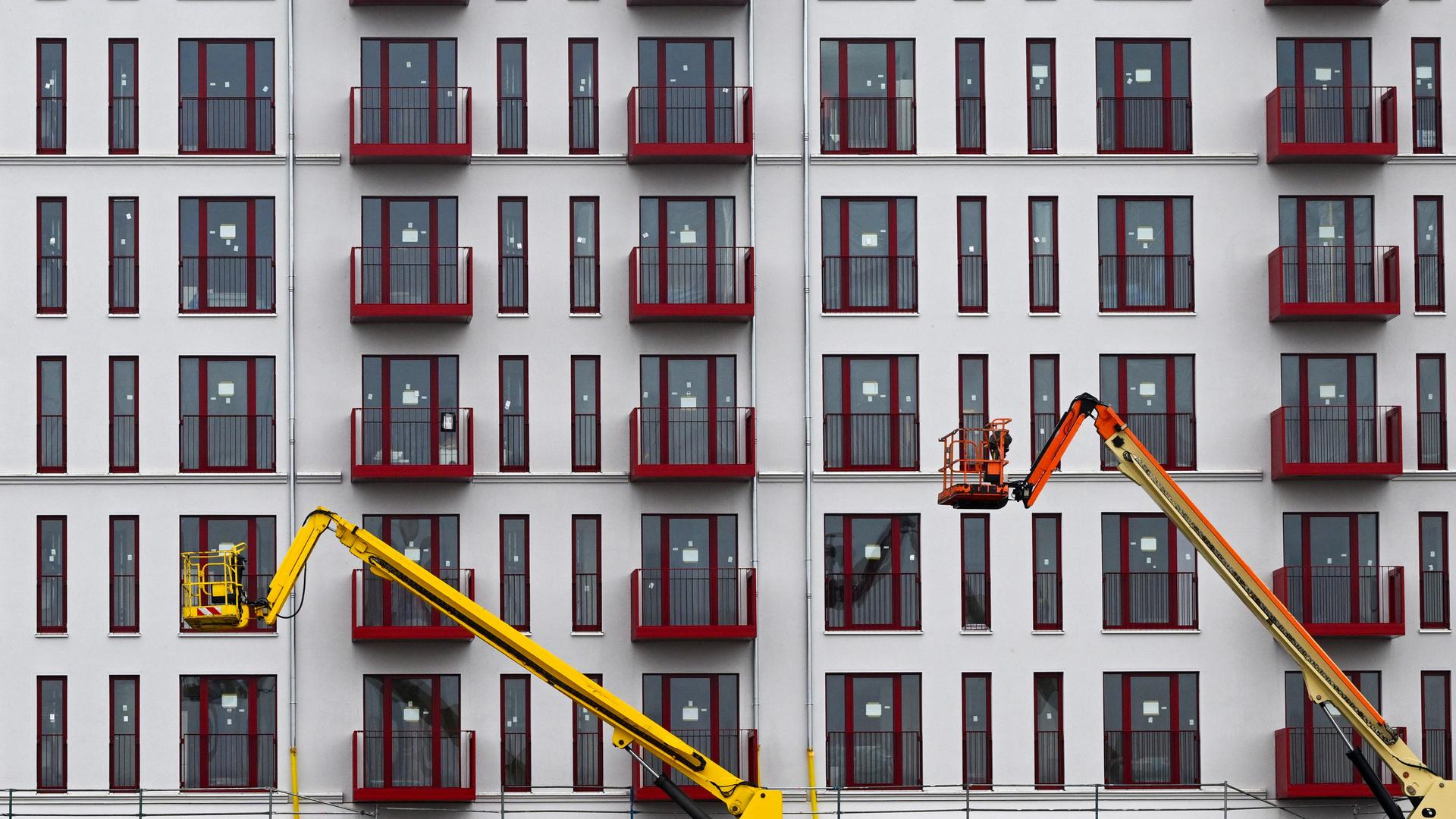 Berlin: An der Fassade eines im Bau befindlichen Mehrfamilienhauses sind zahlreiche Balkone und Fenster zu sehen, zwei Hubarbeitsbühnen stehen vor dem Gebäude. 