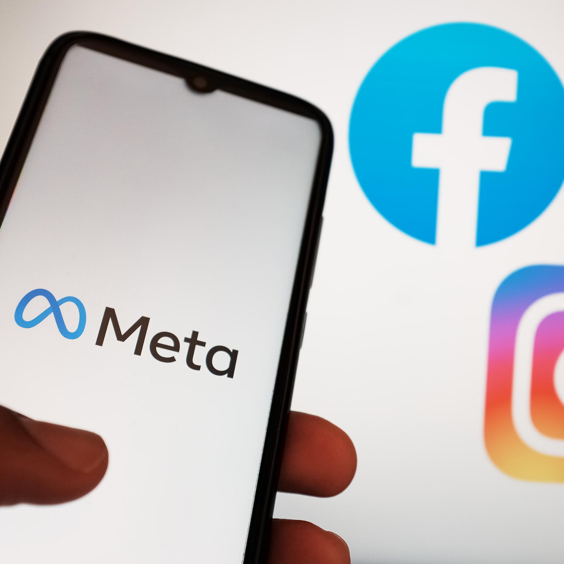 Auf dem Foto sieht man eine Hand. Sie hält ein Handy. Auf dem Bild-Schirm sieht man das Logo von der Firma Meta. Daneben sieht man die Logos von Instagram und Facebook.