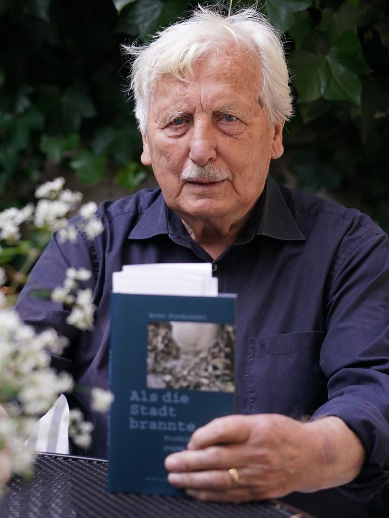 Der Schriftsteller und sein Werk: Arno Surminski mit dem Erzählband "Als die Stadt brannte". Er hält das Cover des Buches Richtung Kamera, um ihn herum sind die Blüten eines Busches zu sehen. 