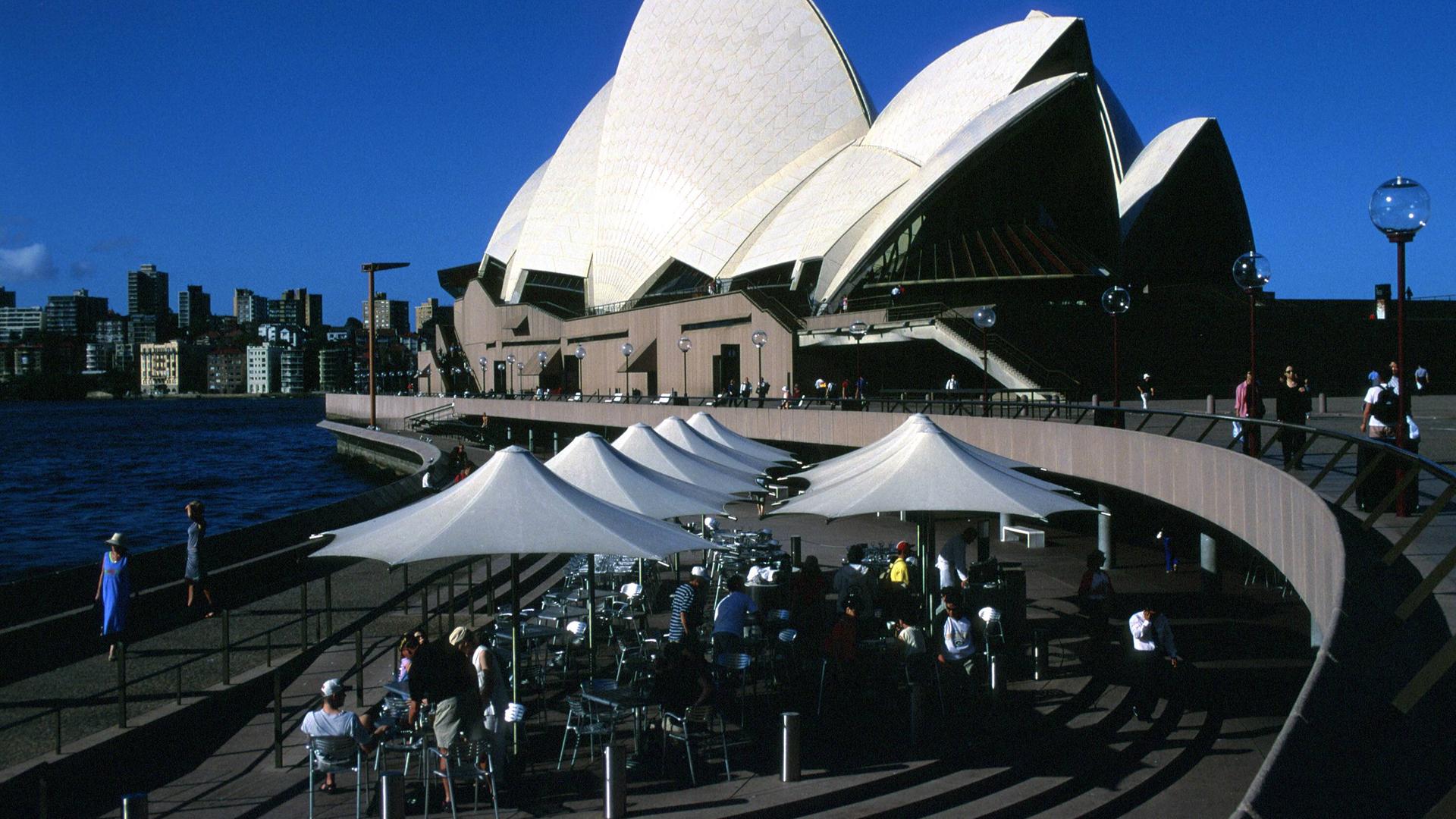 Vor dem Opernhaus in Sydney sitzen Gäste in einem Café auf der Terrasse.