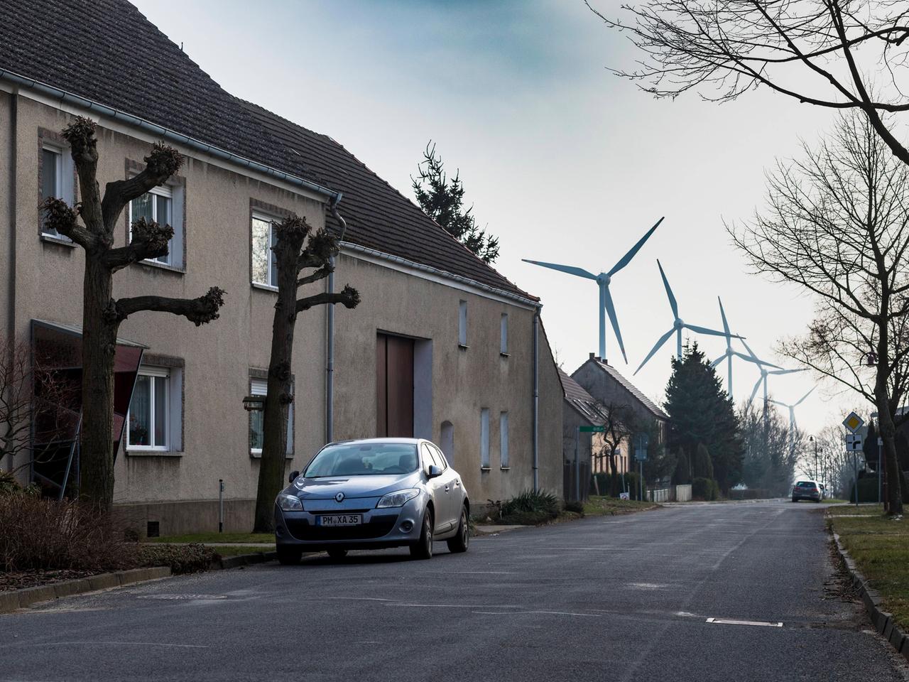 Eine Straße mit Wohnhäusern, Bäumen und einen Auto. Im Hintergrund sind mehrere Windkraftanlagen zu sehen. Es handelt sich um den Ort Feldheim im brandenburgischen Fläming, der sich selbst aus erneuerbarer Energie autark versorgt.