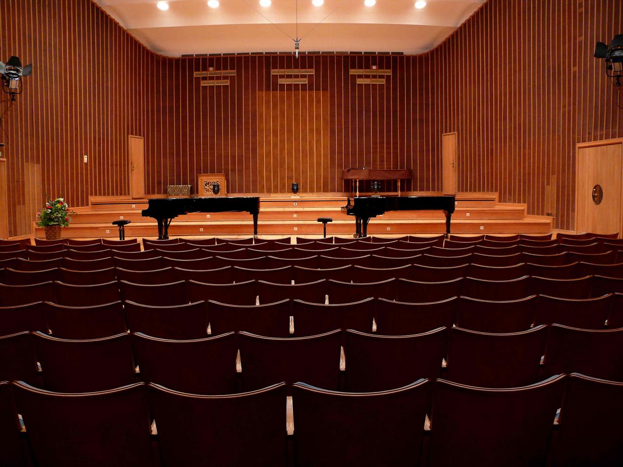 Ein holzgetäfelter Saal ist in warmes Licht getaucht. Vor der Bühne stehen zwei schwarz glänzende Flügel. Im Hintergrund ist ein abgedecktes Cembalo zu sehen. Die Stuhlreihen sind im Saal sind leer. Von der Decke hängt ein Mikrofon.