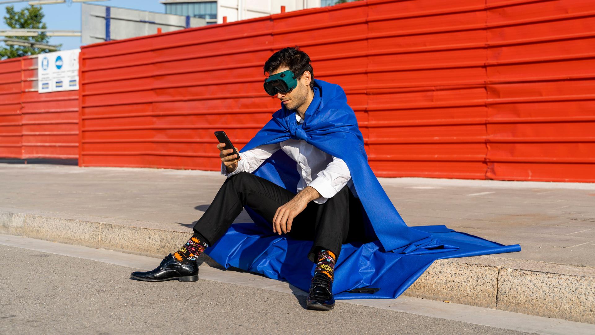 Mann in Anzug und Superhelden-Umhang und -Maske sitzt auf einem Gehsteig und schaut auf sein Smartphone.