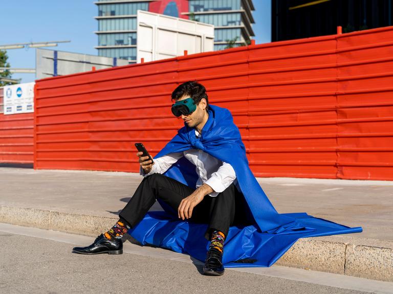 Mann in Anzug und Superhelden-Umhang und -Maske sitzt auf einem Gehsteig und schaut auf sein Smartphone.