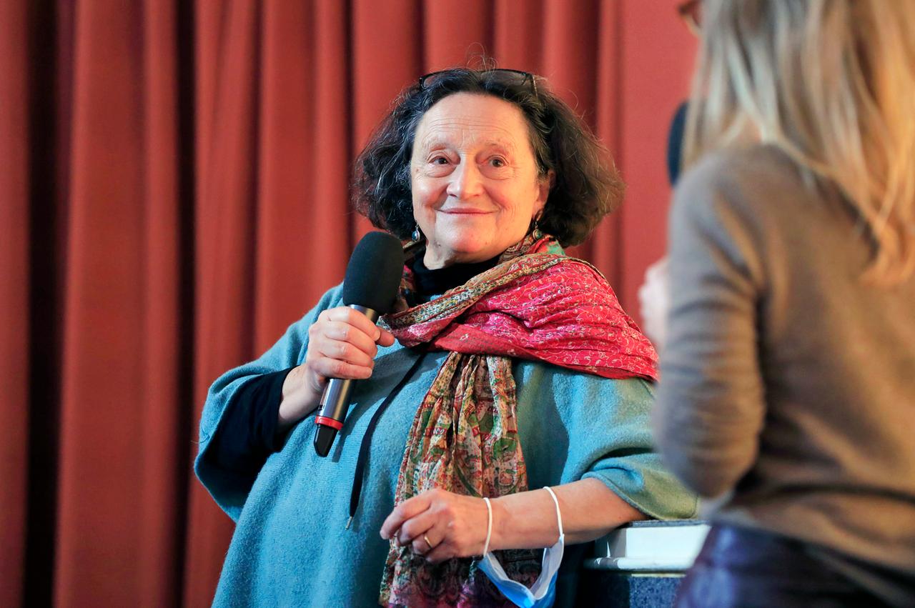 Eine ältere Frau schaut saft, mit einem Lächeln auf den Lippen zu einer angeschnittenen Person am rechten Bildrand und hält ein Mikrofon in der Hand.