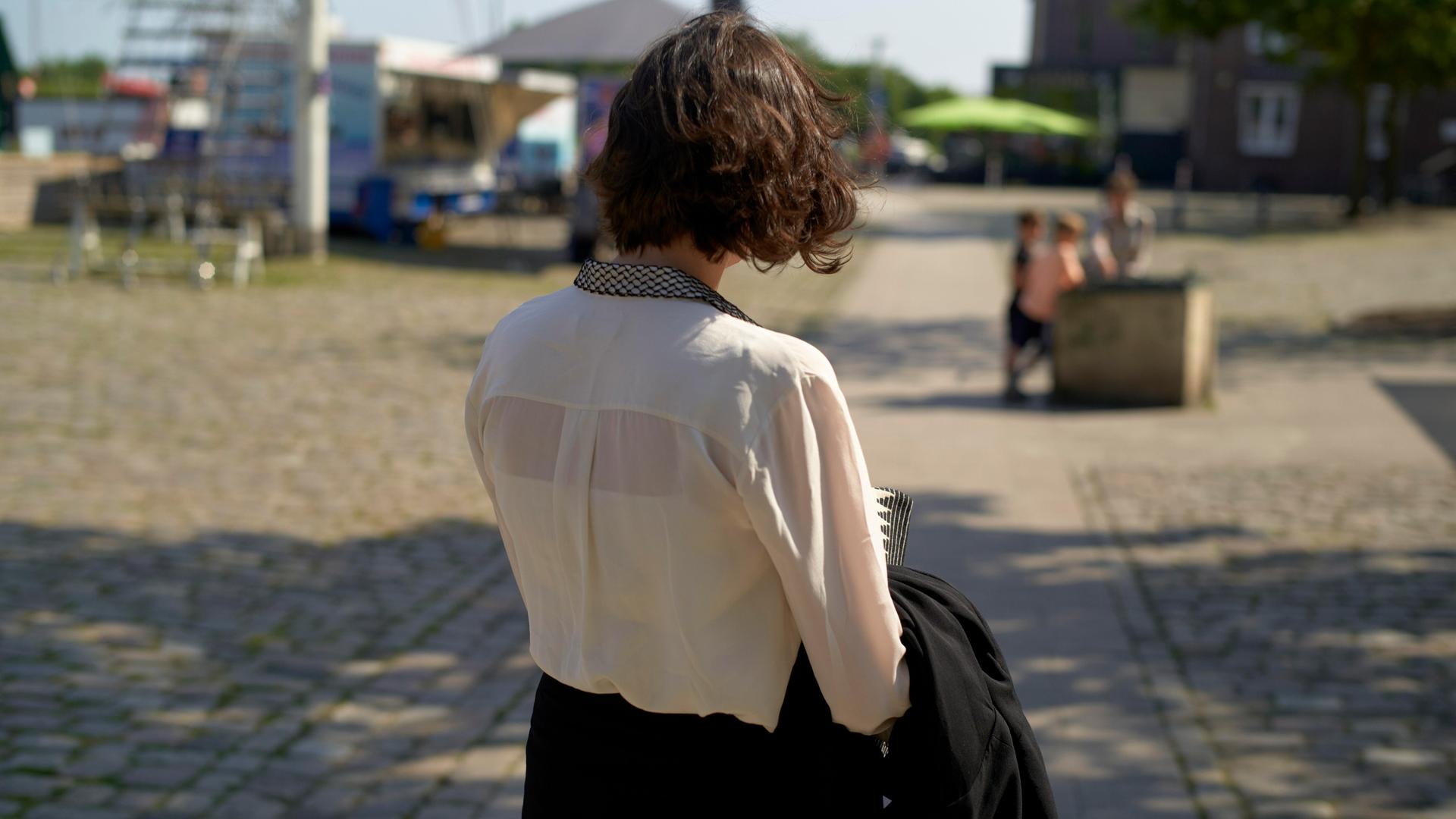 Eine junge Frau mit kurzen braunen Haaren läuft durch eine Stadt bei Sonnenschein und wird von hinten gezeigt
