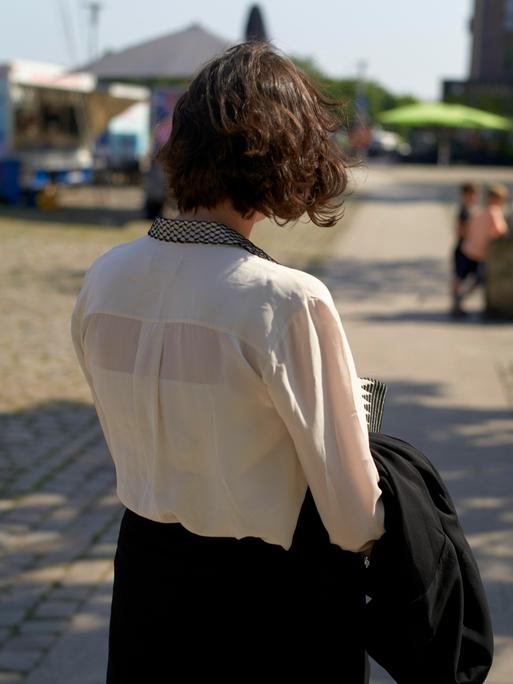 Eine junge Frau mit kurzen braunen Haaren läuft durch eine Stadt bei Sonnenschein und wird von hinten gezeigt