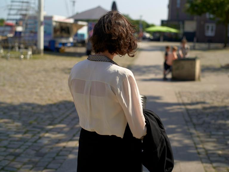 Eine junge Frau mit kurzen braunen Haaren läuft durch eine Stadt bei Sonnenschein und wird von hinten gezeigt