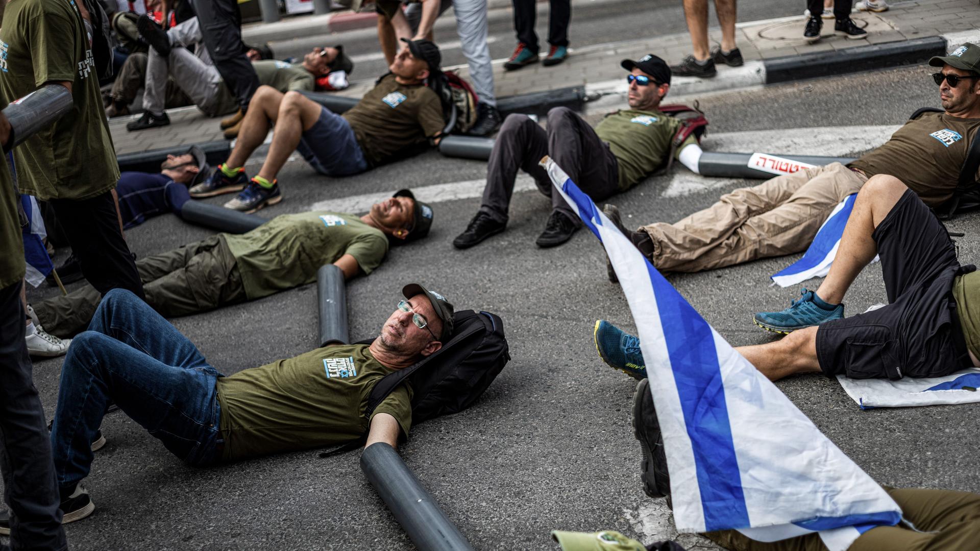 Die Militärreservisten liegen auf dem Boden einer Einfahrt und haben ihre Hände in schwarze Plastikrohre gesteckt. Daneben eine israelische Flagge. Die Militärreservisten liegen auf dem Boden einer Einfahrt und haben ihre Hände in schwarze Plastikrohre gesteckt. Daneben eine israelische Flagge.