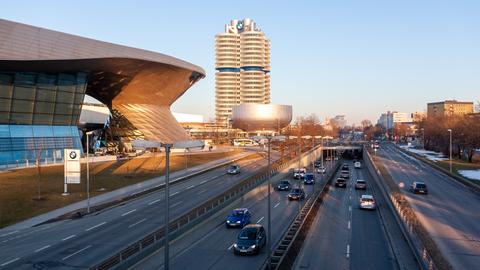 Autos befahren eine Straße, im Hintergrund ist das Hauptgebäude von BMW zu sehen, dessen Architektur an einen Vierzylindermotor angelehnt ist.