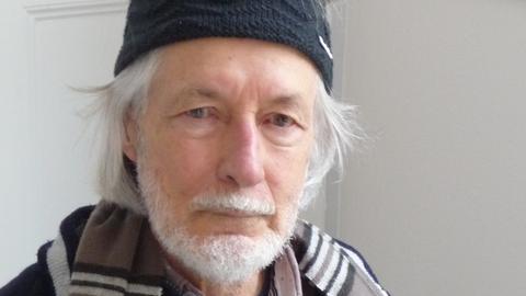 Ein älterer Mann mit Bart grauen Haaren blickt mit Schal und Wollstirnband in die Kamera