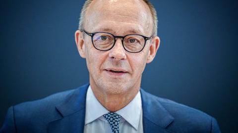 Friedrich Merz schaut in die Kamera. Er trägt eine runde schwarze Hornbrille, einen mittelblauen Anzug und eine blaue Krawatte.