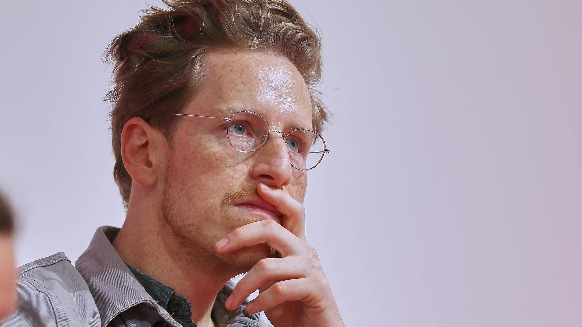 Porträt von Leon Engler mit Hemd und Brille. Seine linke Hand hält er nachdenklich vor dem Mund. Sein Blick ist konzentriert.