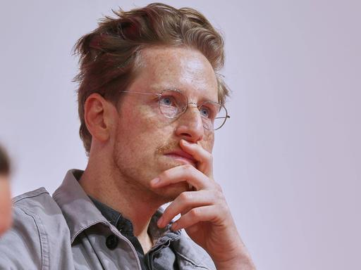 Porträt von Leon Engler mit Hemd und Brille. Seine linke Hand hält er nachdenklich vor dem Mund. Sein Blick ist konzentriert.