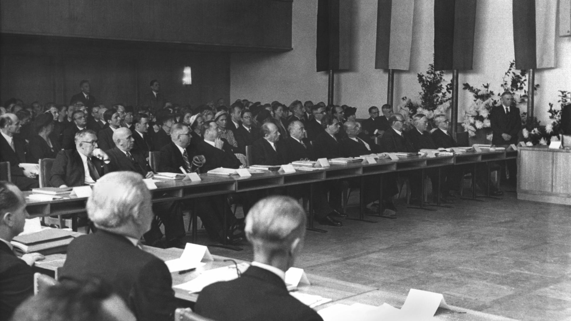 Die Ratsmitglieder sitzen an einem langen, L-förmigen Tisch. Weitere Menschen sitzen auf Stühlen dahinter. Im Hintergrund sind Teile der Flaggen der Bundesländer zu sehen. 