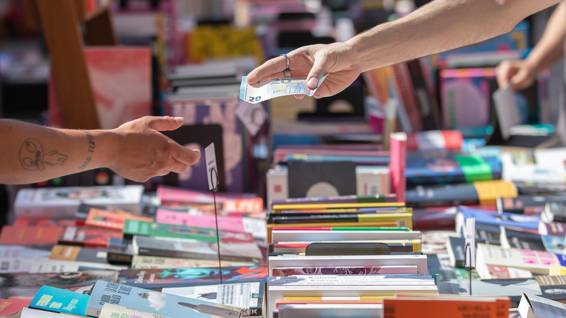 Über einen Tisch voll mit Bücher wird ein 20-Euro-Schein zum Bezahlen gereicht. Über einen Tisch voll mit Bücher wird ein 20-Euro-Schein zum Bezahlen gereicht.
