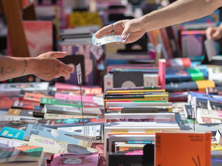 Über einen Tisch voll mit Bücher wird ein 20-Euro-Schein zum Bezahlen gereicht. Über einen Tisch voll mit Bücher wird ein 20-Euro-Schein zum Bezahlen gereicht.